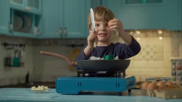 Ung förskola pojke matlagning äggröra själv står ensam på blått kök hemma. Kid bryta ägg med kniv i varm stekpanna på bärbar gasspis. — Stockvideo