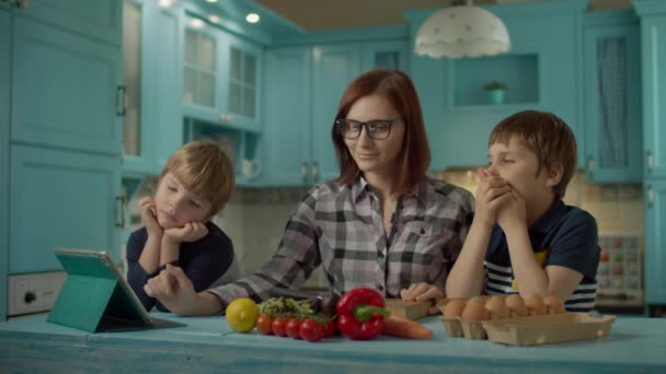 Familie koken samen op zoek naar online recept op tablet computer staan op blauwe keuken. Twee kinderen helpen moeder om voedsel thuis te koken. — Stockvideo