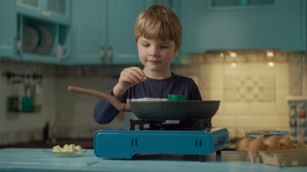 Der kleine Vorschuljunge kocht allein Rührei und steht zu Hause allein in der blauen Küche. Kind legt Butter in heiße Pfanne auf tragbarem Gasherd. — Stockvideo