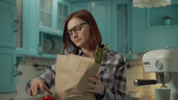 Mulher adulta jovem em óculos segurando saco de papel com alimentos frescos e colocando vegetais e ovos em pé na cozinha azul em casa. Feminino coloca na mesa espargos e outros alimentos. Fechar — Vídeo de Stock