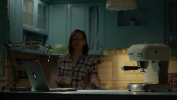 Młoda kobieta po trzydziestce w okularach, pracująca w nocy na laptopie w niebieskiej kuchni. Kobieta biorąc filiżankę i robi kawę podczas pracy na laptopie w domu. — Wideo stockowe