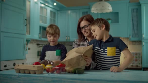 Dos niños ayudan a la joven madre de 30 años a desempacar la bolsa de papel con alimentos frescos. Niños y mujeres que ponen verduras, huevos y pasta en la mesa de la cocina en casa. Cocina familiar juntos . — Vídeos de Stock