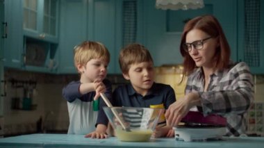 Evdeki mavi mutfakta waffle makinesinde waffle yapan iki çocuklu genç bir anne. Çocuk kaşıkla waffle makinesine hamur koyuyor. Birlikte yemek pişirmek..