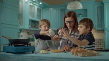 Mutlu anne ve oğullu bir aile evdeki mavi mutfakta omlet pişiriyor. Çocuklar yumurtayla cam kaseye tuz ve biber koyuyorlar. Taşınabilir gaz ocağında kızartma tavası. 