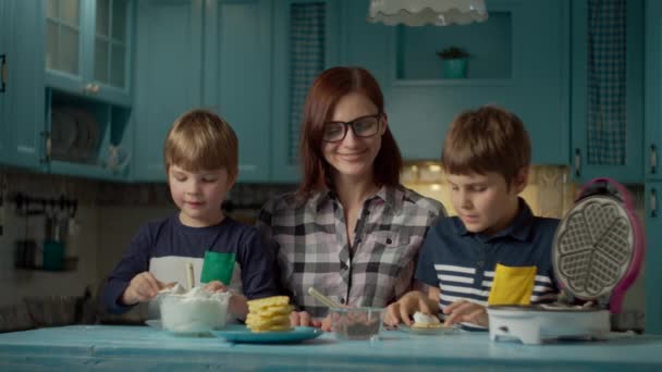 30代の若い母親は、ブルーキッチンに立って自家製ワッフルにクリームとチョコレートを置くために2人の子供を助けます。子供たちは自作のお菓子を食べることを楽しむ。一緒に料理する. — ストック動画