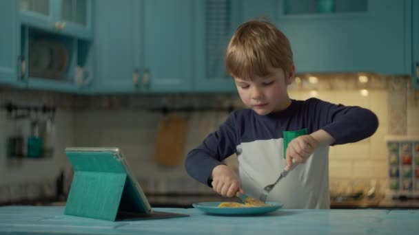 Niño comiendo huevos revueltos con tomates viendo tableta en casa. Niño comiendo desayuno hecho a sí mismo con gadget en casa . — Vídeo de stock