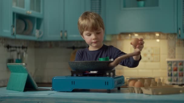 Niño preescolar cocinar huevos revueltos mirando receta en línea en la computadora de la tableta de pie solo en la cocina azul en casa. Niño rompiendo huevos con cuchillo en sartén caliente en estufa de gas portátil . — Vídeos de Stock
