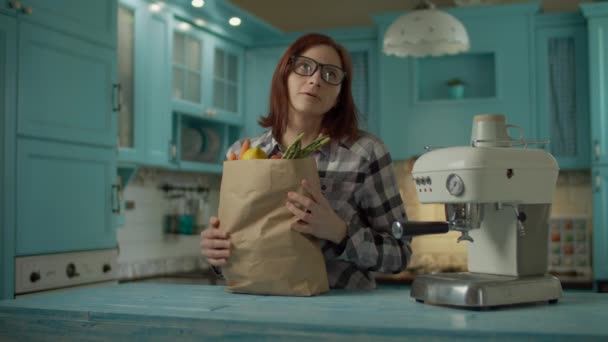 Mujer joven adulta en gafas cansada de sostener la bolsa de papel con comida fresca comienza a poner las verduras de pie en la cocina azul en casa. Las mujeres encienden la máquina de café para hacer una taza de café . — Vídeos de Stock