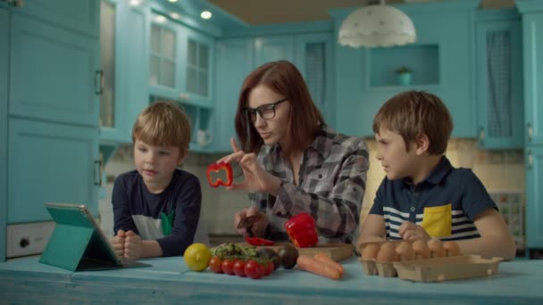 Familia cocinar juntos mirando receta en línea en la computadora de la tableta de pie en la cocina azul. Dos niños ayudando a mamá a cocinar en casa. Mujer rebanando pimiento rojo dulce . — Vídeos de Stock