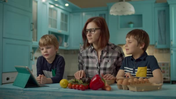 Die Familie kocht zusammen und betrachtet das Online-Rezept auf dem Tablet-Computer, der auf der blauen Küche steht. Zwei Kinder helfen Mama beim Kochen zu Hause. Frau schneidet Paprika in Scheiben. — Stockvideo