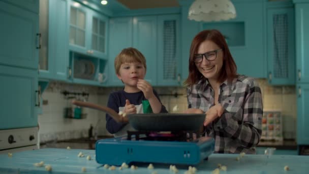 Joyeux famille de la jeune mère et son fils cuisson pop maïs sur poêle à frire sur cuisine bleue à la maison. Explosions de maïs sur poêle chaude au ralenti. Enfant manger du maïs soufflé pendant la cuisson — Video