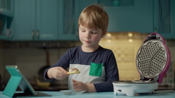 취학 전 남자 아이가 아이스크림 과 초콜릿으로 집에서 와플을 먹으면서 파란 부엌에 서 있는 태블릿 컴퓨터를 보고 있습니다. 아이들은 스스로 만든 사탕을 먹고 기기를 보는 것을 즐긴다. — 비디오