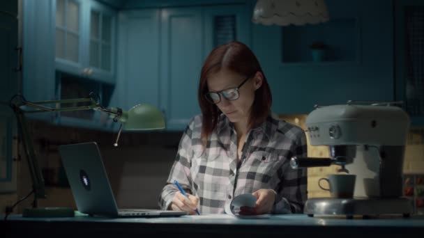 Mujer joven de 30 años en gafas que trabajan en el ordenador portátil desde casa en la cocina azul por la noche. Mujer de negocios firmando documentos en papel trabajando desde casa . — Vídeos de Stock