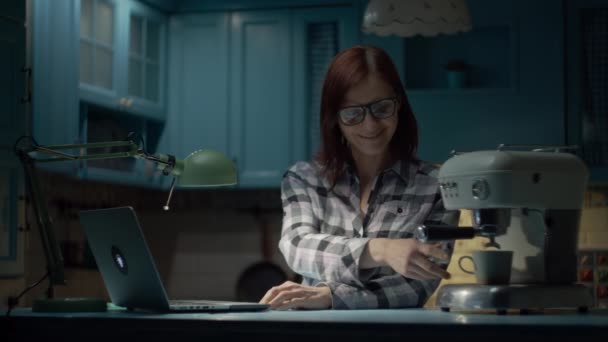 Jonge vrouw van in de 30 met een bril die thuis op een laptop werkt in een blauwe keuken. Vrouwelijke drinken kopje koffie tijdens het werken op laptop thuis 's nachts. — Stockvideo