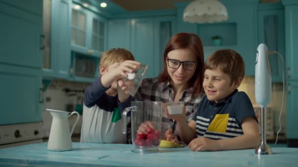 母と2人の子供の幸せな家族は、ホームブルーキッチンでミキサーでイチゴ、バナナ、ビーガンミルク、ヨーグルトのスムージーを調理します。イチゴとヨーグルトをボウルに注ぐ男の子. — ストック動画