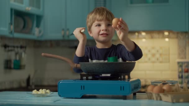 Az óvodás fiú rántottát főz, egyedül áll otthon a kék konyhán. Gyerek feltöri a tojást késsel forró serpenyőbe a hordozható gáztűzhelyen. — Stock videók
