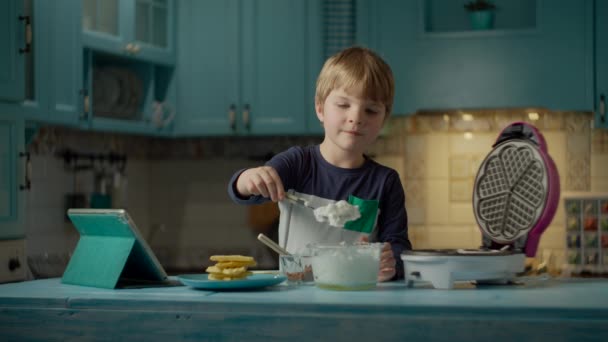 Menino pré-escolar comendo waffles caseiros com creme e chocolate, assistindo tablet computador em pé na cozinha azul. Kid gosta de comer doces auto-feitos e assistir gadget . — Vídeo de Stock