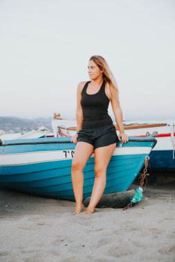 Sahilde eski bir mavi teknesinde oturan sarışın kız.