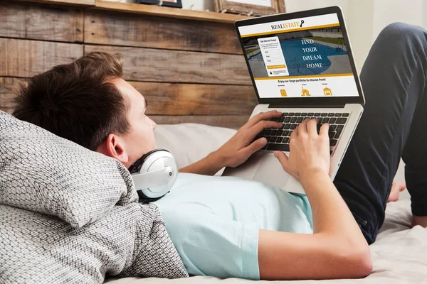 Man zoekt huis in een onroerend goed website met een laptop liggend op het bed. — Stockfoto