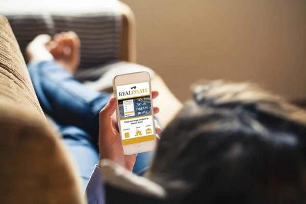 Woman searching for house through mobile phone in a real estate app while lying on the sofa at home. — Stock Photo, Image