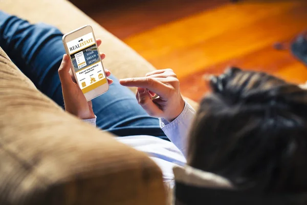 Mujer usando el teléfono inteligente para buscar casa en un sitio web de bienes raíces mientras está acostado en casa . — Foto de Stock