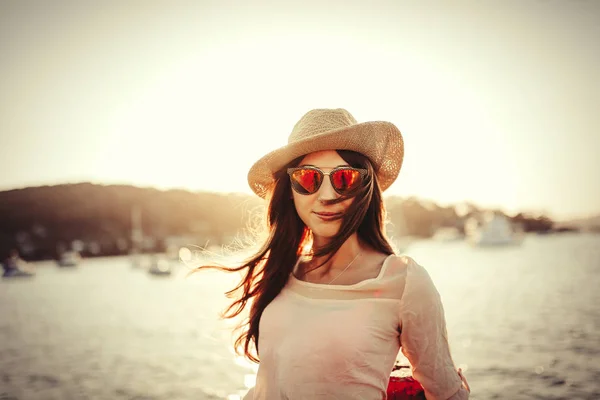 Frau mit Hut und Sonnenbrille posiert bei Sonnenuntergang mit dem Meer im Hintergrund. — Stockfoto