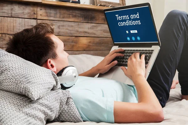 Man die termen en voorwaarden op een website met een laptop leest terwijl liggend op het bed thuis. — Stockfoto
