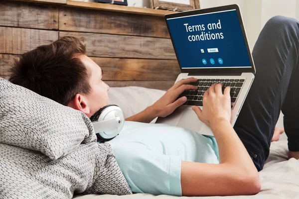 Hombre leyendo Términos y condiciones en un sitio web con un ordenador portátil mientras está acostado en la cama en casa . — Foto de Stock