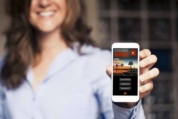 Mujer sonriente sosteniendo un teléfono móvil en la mano con la aplicación de streaming de vídeo en la pantalla . — Foto de Stock