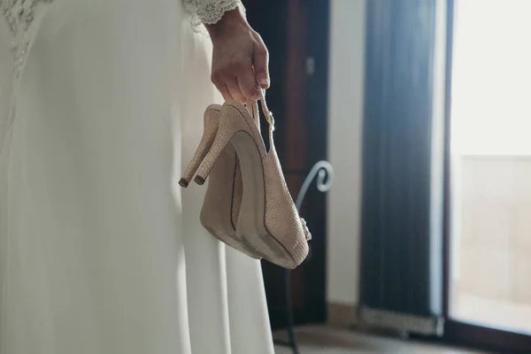 Невеста держит пару свадебных туфель в руке, перед окном — стоковое фото