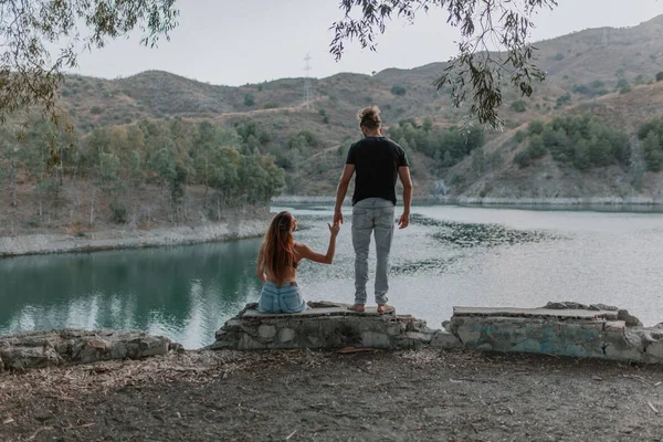 Hombre sosteniendo la mano de mujer mientras ella está sentada en la orilla del río en la naturaleza — Foto de Stock