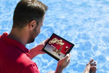 Adam havuz üzerinde otururken Noel hediyeleri elektronik bir tablet ile internet alışveriş.