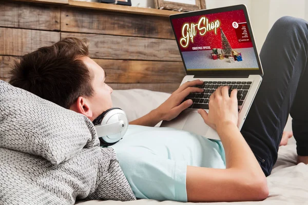 Ο άνθρωπος ψώνια χριστουγεννιάτικα δώρα με ένα φορητό υπολογιστή μέσω internet ενώ ξεκούραση στο κρεβάτι στο σπίτι. — Φωτογραφία Αρχείου