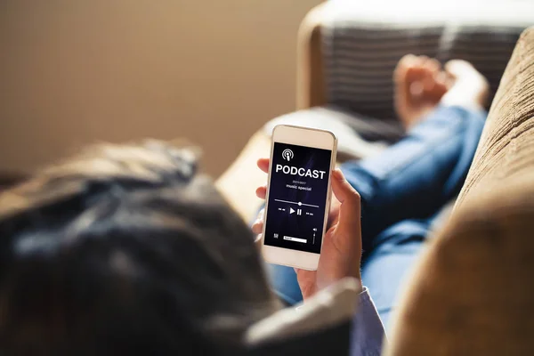 Donna a casa con in mano un telefono cellulare con app podcast sullo schermo mentre si sdraia sul divano . — Foto Stock
