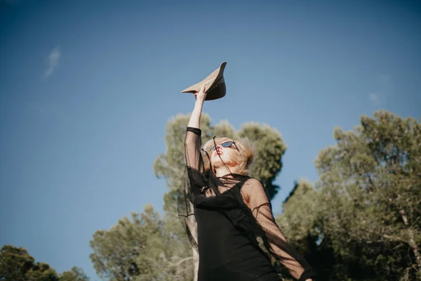 Jonge blonde vrouw springen in de natuur met zwarte zonnebrillen en kleding en een hoed. — Stockfoto