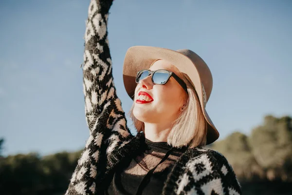 Beautiful blonde girl with sunglasses, hat and red lips enjoying outdoors, while raising arm up. — Stock Photo, Image