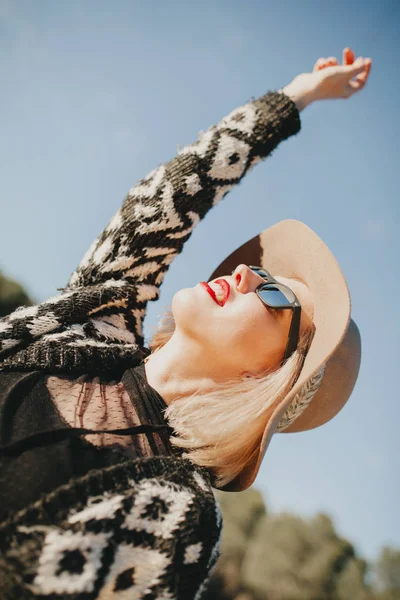 Beautiful blonde girl with sunglasses, hat and red lips enjoying outdoors, while raising arm up. — Stock Photo, Image