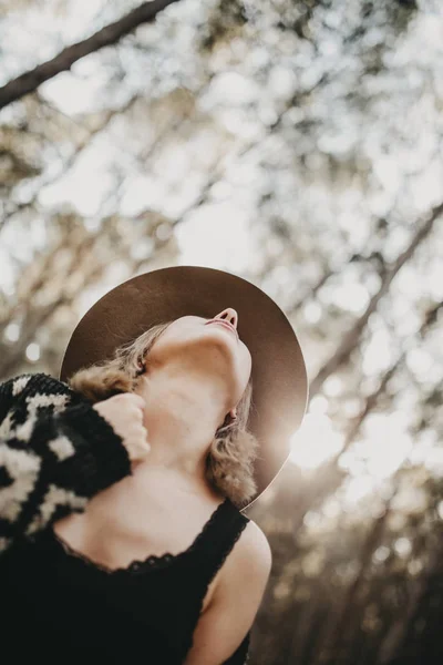 Blonde woman with a hat looking up in the countryside. Forest trees in the background. — Stock Photo, Image