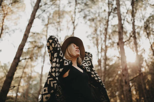 Blonde woman with a hat looking up in the countryside. Forest trees in the background. — Stock Photo, Image