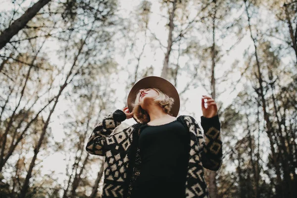 Blonde woman with a hat looking up in the countryside. Forest trees in the background. — Stock Photo, Image