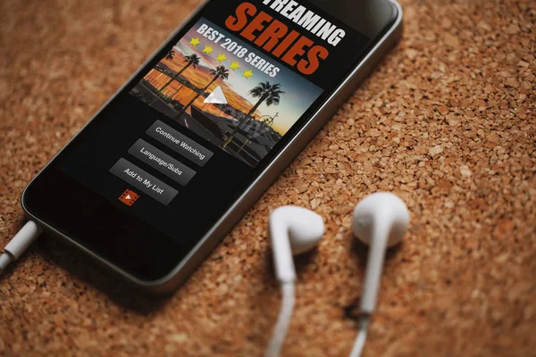 Close up af streaming serie app i en mobiltelefon skærm, tæt på hvide øretelefoner på en brun bord . - Stock-foto