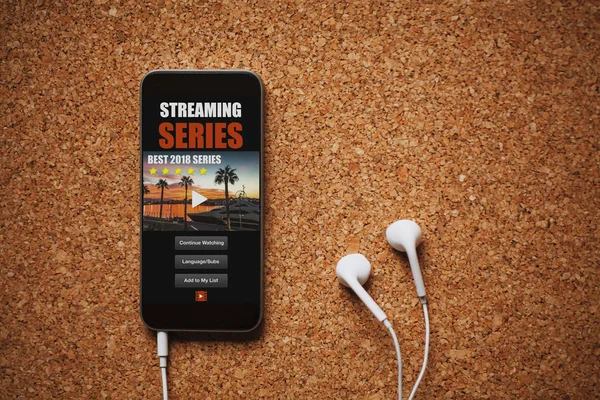 Frontansicht der Streaming-Serien-App auf einem Handy-Bildschirm, in der Nähe weißer Kopfhörer auf einem braunen Tisch. — Stockfoto