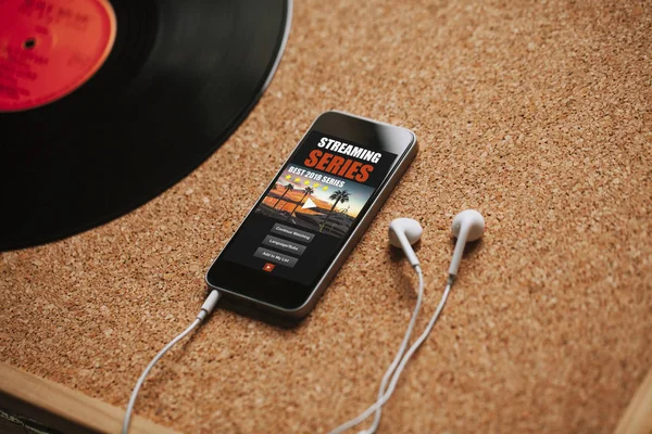 Streaming series app in a mobile phone screen, close to white earphones on a brown table.