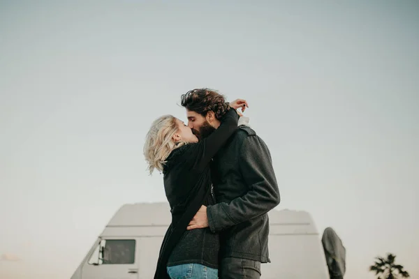 Cool jovem casal beijando uns aos outros ao ar livre, enquanto eles são abraçados, durante uma parada de viagem, com sua van no fundo . — Fotografia de Stock