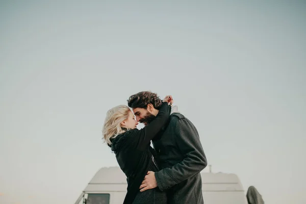 Cool pareja joven besándose al aire libre mientras son abrazados, durante una parada de viaje por carretera, con su furgoneta en el fondo . — Foto de Stock