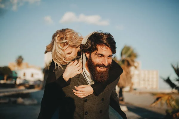 Cool indie casal se divertindo ao ar livre, enquanto ele lhe dá um piggyback . — Fotografia de Stock