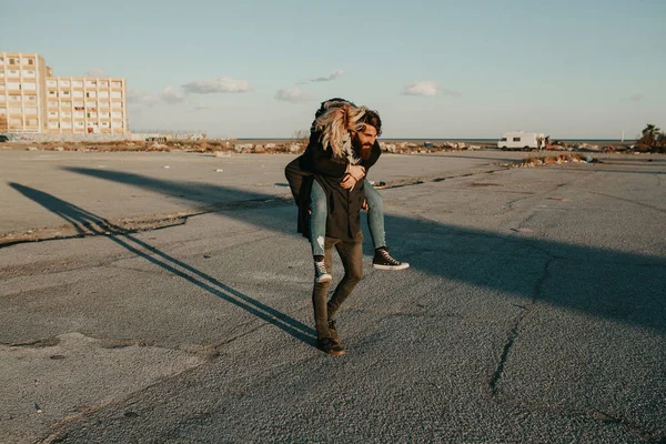 Cool indie casal se divertindo ao ar livre, enquanto ele lhe dá um piggyback em um lugar abandonado . — Fotografia de Stock