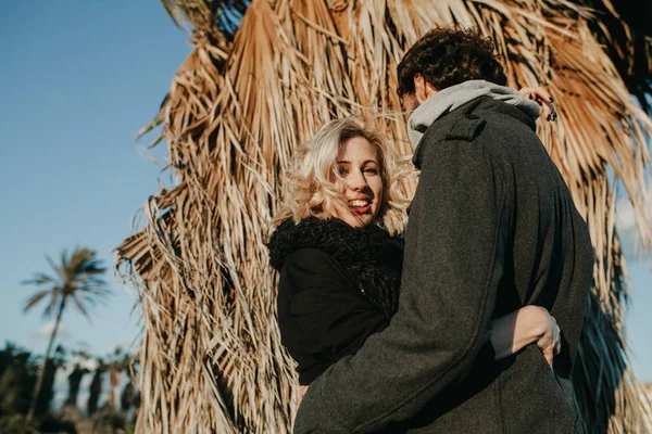 Mulher loira sorrindo enquanto ela abraça seu namorado ao pôr do sol com uma palmeira no fundo . — Fotografia de Stock