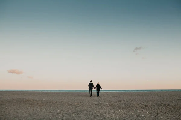 Чоловік і жінка, що йде на пляжний пісок на заході сонця, тримаючись за руки, з морем і блакитним небом на задньому плані . — стокове фото