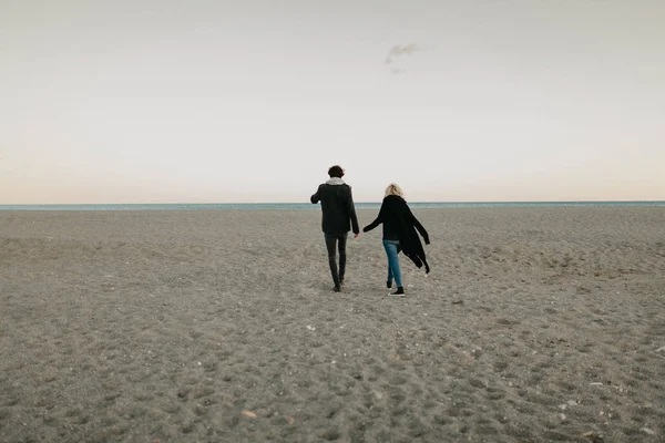 Pria dan wanita berjalan di pantai pasir saat matahari terbenam, berpegangan tangan, dengan laut dan langit matahari terbenam di latar belakang . — Stok Foto